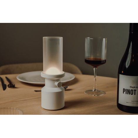Lampe de table blanche allumée posée sur une table dressée avec assiette, verres et bouteille, sans marquage visible.