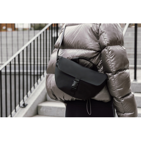 Sac bandoulière noir porté en travers du buste par une personne en extérieur, sans marquage visible.