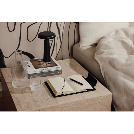 Carnet de notes noir ouvert posé sur une table de chevet avec stylo, verres et lampe, sans marquage visible.