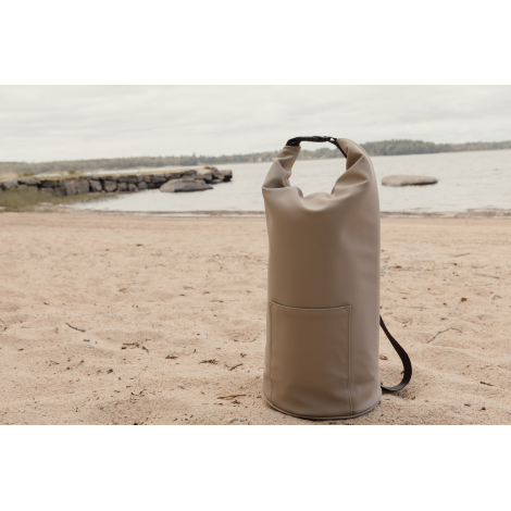 Sac isotherme roll-top beige posé sur le sable près de l’eau avec poche frontale, sans marquage visible.