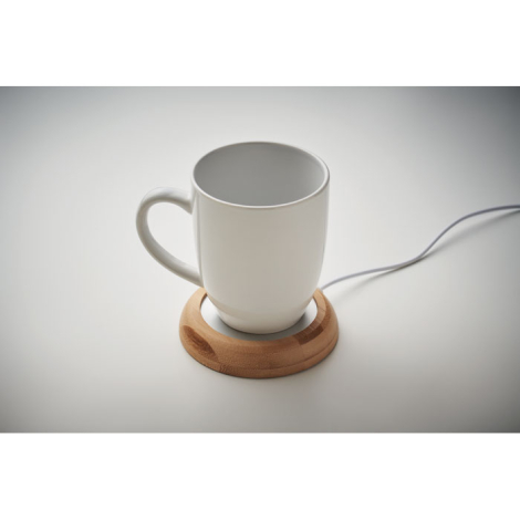 Chauffe-tasse rond en bambou avec tasse blanche posée dessus, câble branché, sans marquage.