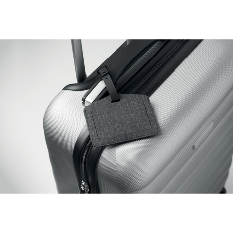 Étiquette de bagage en tissu gris chiné fixée sur une valise rigide argentée, sans marquage.