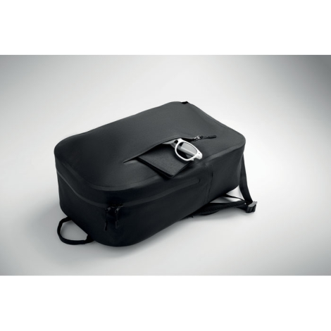 Sac à dos noir en textile vu de dos posé à plat, poche arrière ouverte laissant apparaître une paire de lunettes, sans marquage.