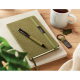 Carnet vert rigide avec élastique assorti, stylo et porte-clés posés sur une table avec tasse et plantes, marquage visible.