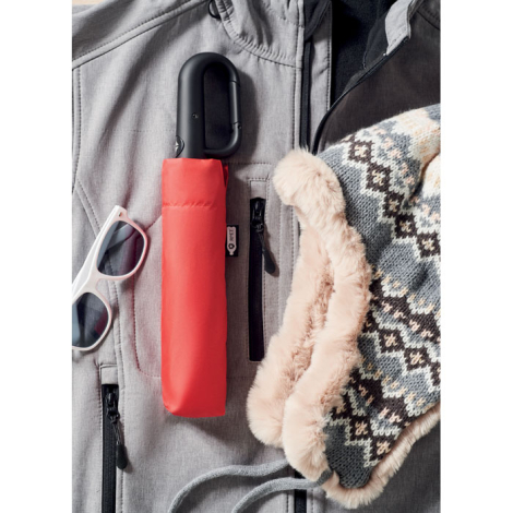 Parapluie rouge plié dans sa housse assortie posé sur vêtement gris avec lunettes et bonnet, sans personnalisation visible.