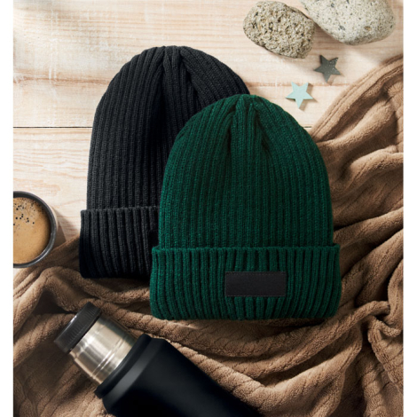 Deux bonnets tricotés noir et vert posés sur une surface en bois avec objets du quotidien, sans personnalisation visible.