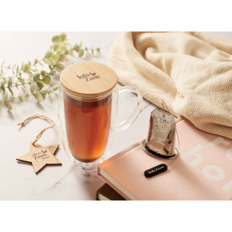 Mug en verre double paroi rempli de thé, couvercle en bambou gravé et accessoires posés autour, marquage visible sur le couvercl