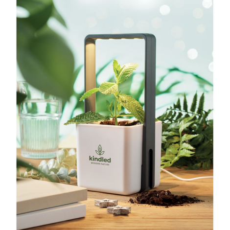 Pot de culture blanc posé sur une table, contenant une plante verte sous arceau LED noir, marquage visible sur la face avant.