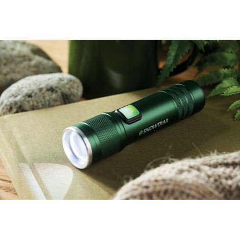 Lampe torche en aluminium vert foncé posée sur une surface textile beige avec pierres et feuillage, marquage visible sur la face