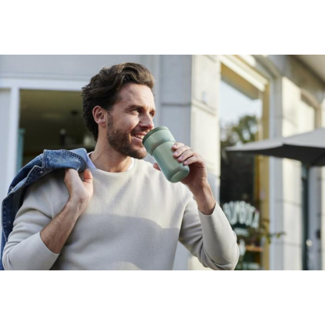 Homme tenant un mug de voyage Ellipse vert en extérieur et buvant, marquage visible sur la partie centrale.