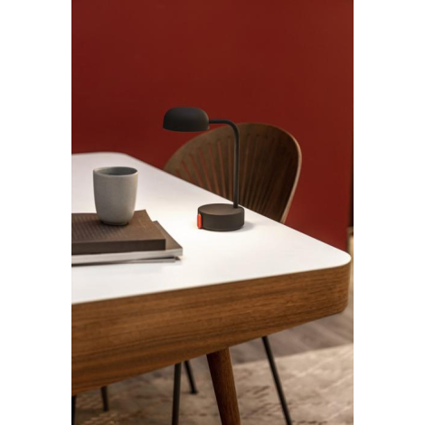 Lampe noire posée sur une table en bois clair, à côté d’une tasse et d’un carnet, sans marquage visible.