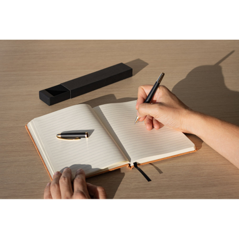 Roller noir et or utilisé sur un carnet ouvert, capuchon posé sur la table, sans marquage apparent.