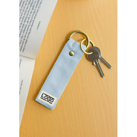 Porte-clés bleu clair posé sur bois clair près d’un livre ouvert et d’un trousseau, marquage face avant.