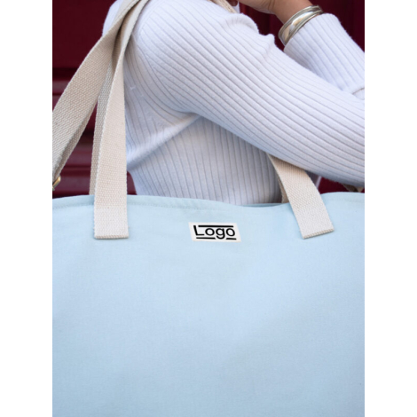 Sac bleu clair porté à l’épaule par une personne vêtue de blanc, marquage visible.