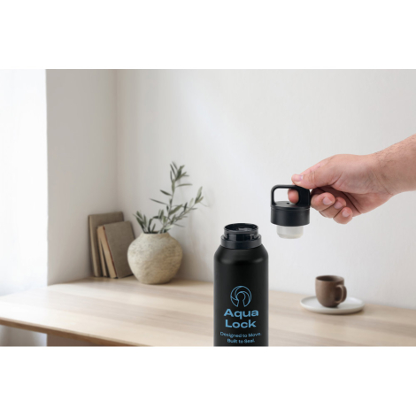 Bouteille isotherme noire avec bouchon dévissé tenu par une main sur une table claire, marquage visible sur la face avant.