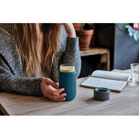 Tasse de voyage utilisée dans un café, tenue en main avec le couvercle posé sur la table, manchon en silicone assurant une isola