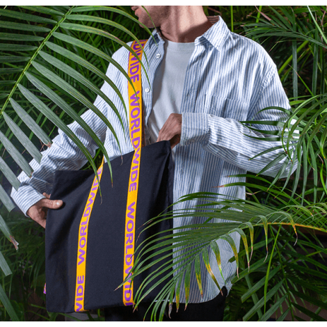 Vue de trois quarts d’un tote bag noir porté par un homme, partiellement masqué par des plantes, avec anses jacquard jaunes et v