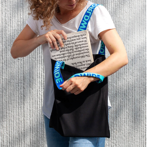 Vue de trois quarts d’un tote bag noir porté par une femme, montrant l’ouverture du sac et les anses jacquard bleues posées sur 