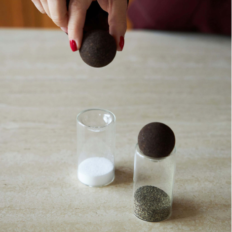 Gros plan d’une main retirant un bouchon sphérique en liège foncé d’un petit pot en verre, tandis qu’un second pot rempli est po