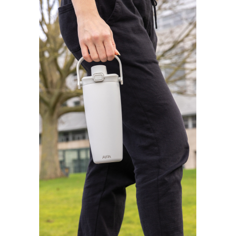 Mug isotherme blanc porté à la main par une personne en extérieur, transporté par la poignée supérieure, sans bandoulière visibl