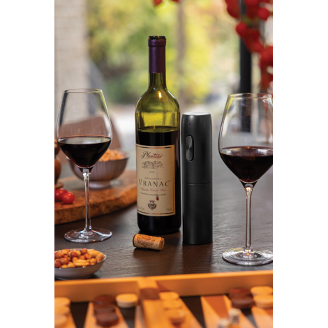 Tire-bouchon électrique positionné verticalement sur une table aux côtés d’une bouteille de vin ouverte, de deux verres remplis 
