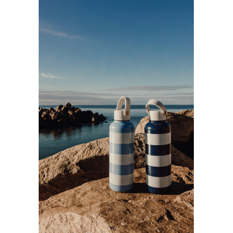 Deux gourdes isothermes identiques en acier inoxydable posées debout sur des rochers face à un plan d’eau, avec motif rayé bleu 