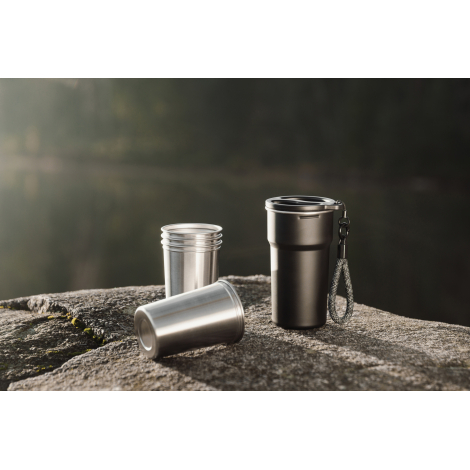 Scène extérieure montrant une tasse de rangement noire avec anse et cordon, accompagnée de trois tasses en acier inoxydable posé
