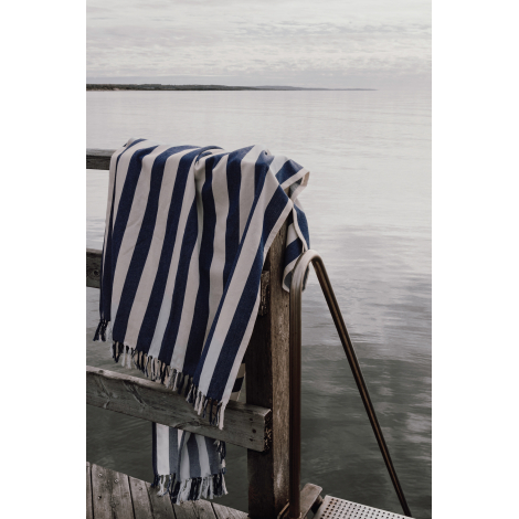 Serviette à rayures bleues et blanches drapée sur une structure en bois au bord de l’eau, avec franges visibles aux extrémités, 