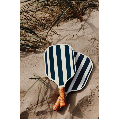 Deux raquettes de pickleball à motif rayé bleu et blanc posées sur le sable avec une balle, dans un environnement extérieur, les