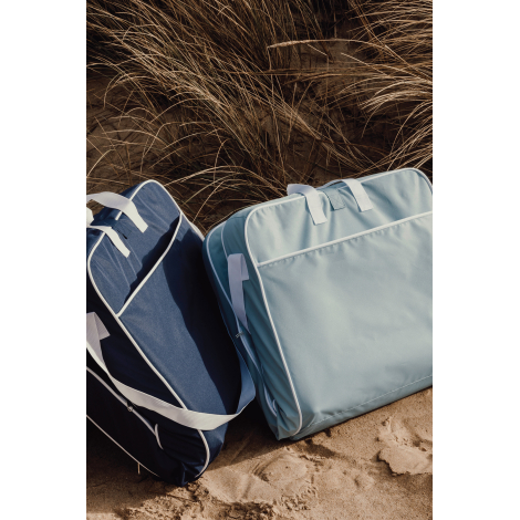 Deux chaises longues pliées posées sur le sable, l’une bleu clair et l’autre bleu foncé, avec poignées et sangles de portage vis