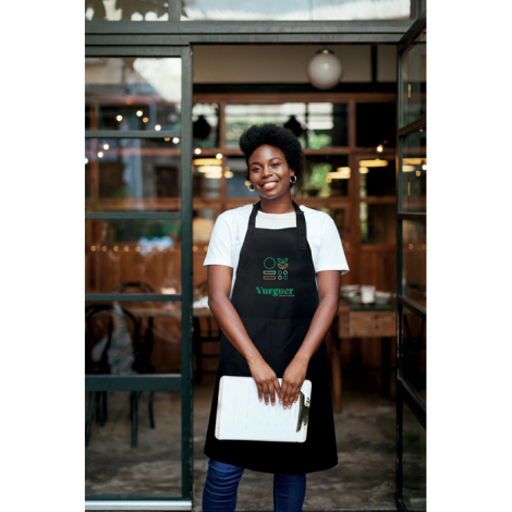 Tablier de cuisine noir porté par une femme en intérieur, vu de face, avec deux poches frontales et sangle de cou visible, la pa