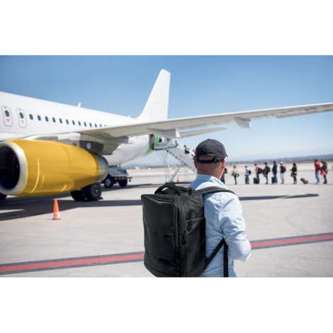 Scène de vie montrant un homme portant un sac à dos cabine sur un tarmac d’aéroport face à un avion, sans marquage visible sur l