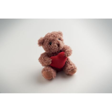 Ours en peluche présenté en légère perspective sur fond clair, assis et tenant un cœur rouge, avec détails brodés du visage visi