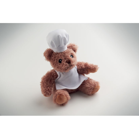Vue en légère perspective d’un ours en peluche chef cuisinier assis sur fond clair, portant une toque et un tablier blancs, sans