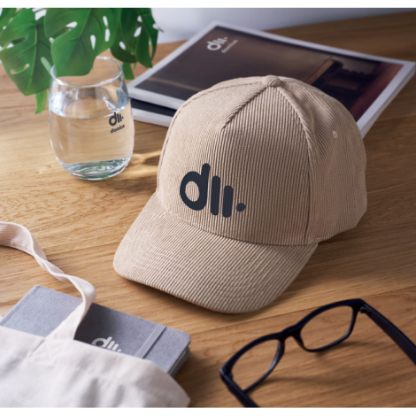 Casquette en velours côtelé beige posée sur une table en bois en intérieur, visière courbée, marquage visible sur le panneau ava
