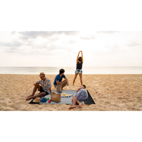 Tapis de plage posé sur le sable en extérieur avec plusieurs personnes assises ou allongées dessus, sans marquage visible.