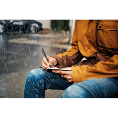Bloc-notes tenu à la main en extérieur sous la pluie pendant l’écriture avec stylo noir visible, sans personnalisation visible.