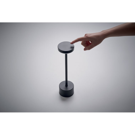 Lampe de table en aluminium sur fond clair avec une main touchant la surface supérieure de la tête pour illustrer la commande ta