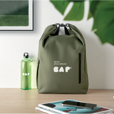 Sac à dos roll top vert posé sur un bureau, vue de face avec marquage visible centré au-dessus de la poche zippée.