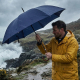 Parapluie bleu foncé ouvert tenu par un homme en extérieur sous la pluie dans un paysage côtier, sans marquage visible sur la to