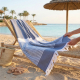 Fouta bleue à rayures disposée sur un transat en bois sur une plage, franges visibles aux extrémités, aucune personnalisation ap