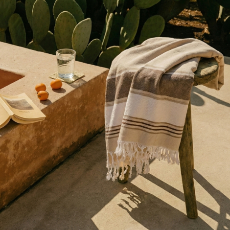 Fouta beige disposée sur un banc en extérieur avec franges visibles et bandes horizontales contrastées, sans marquage apparent.