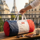 Sac polochon tricolore bleu, blanc et rouge posé sur une table en extérieur, anses blanches et fermeture zippée visibles, marqua