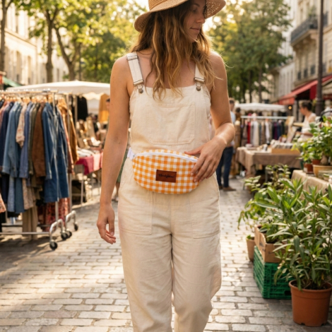 Sac banane à motif vichy orange et blanc porté à la taille par une femme en extérieur, fermeture zippée visible sur le dessus, m