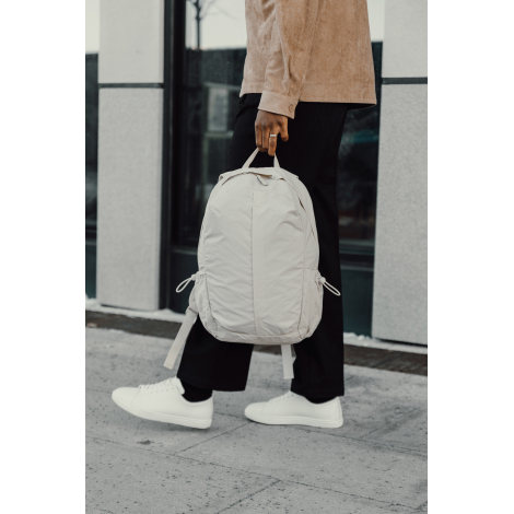 Sac à dos beige porté à la main par une personne en extérieur, sans personnalisation visible