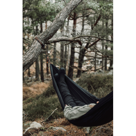 Hamac noir suspendu à un arbre en forêt, avec une personne allongée, sans marquage visible sur le produit.