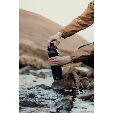 Personne utilisant une gourde en extérieur près d’un cours d’eau, sans marquage visible.