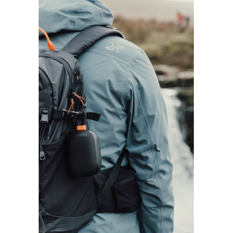 Enceinte accrochée à un sac à dos porté en extérieur, sans personnalisation visible.
