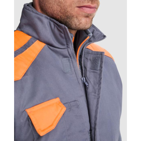Zoom sur le col montant, la fermeture zippée et la poche poitrine à rabat d’un gilet de travail gris avec détails orange, porté 