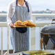 Femme portant un tablier de cuisine à rayures bleu foncé et blanches en tenant un plateau, sans personnalisation visible. Femme portant un tablier de cuisine à rayures bleu foncé et blanches en tenant un plateau, sans personnalisation visible.